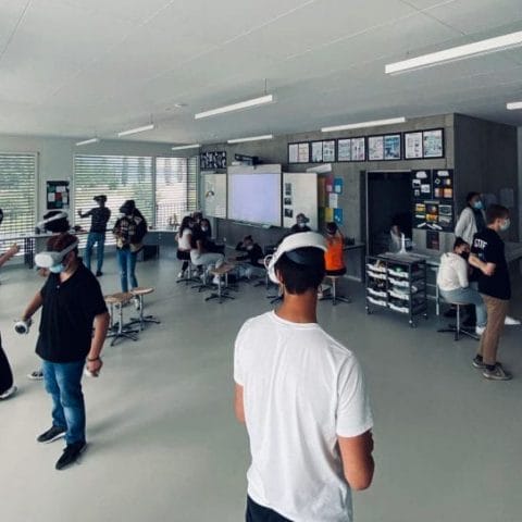 Group of students wearing VR headsets and holding controllers in a spacious classroom, participating in an interactive learning activity.