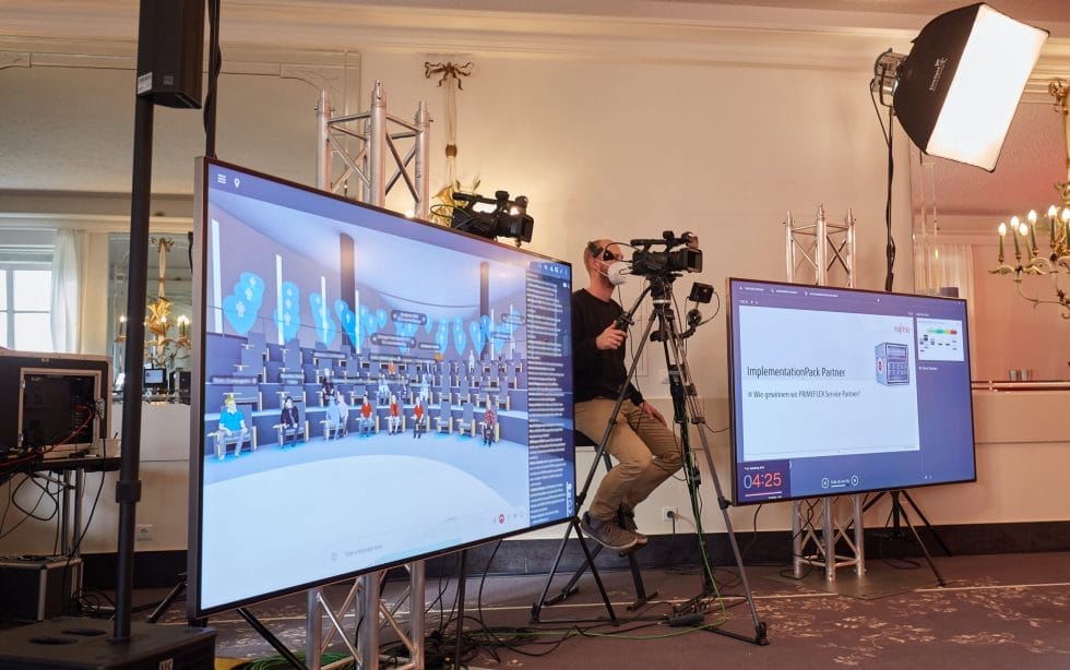 Professional camera crew records a hybrid event with large screens displaying a virtual conference in Hyperspace Metaverse Platform and presentation slides.